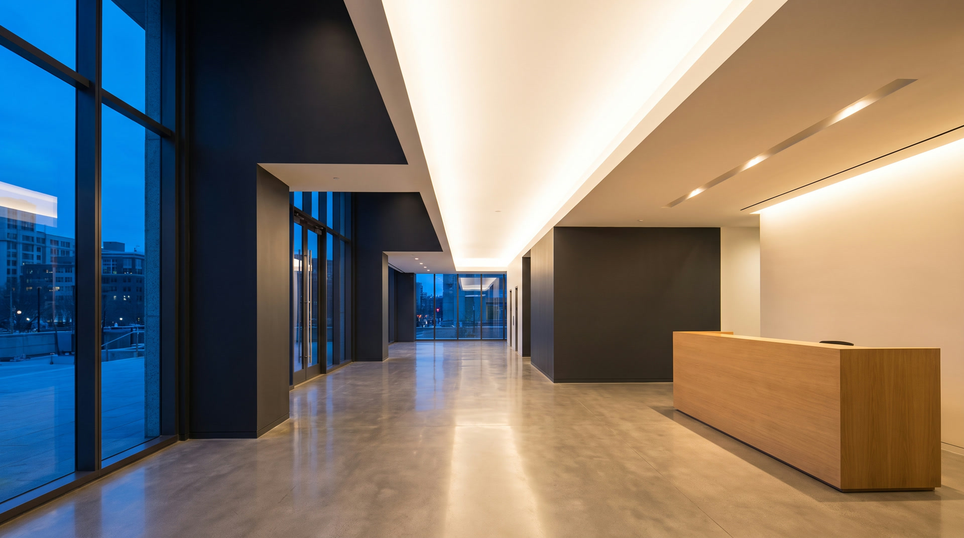 An empty office lobby after hours: charcoal and white walls meeting in sharp lines, an oak reception desk, and blue twilight outside.